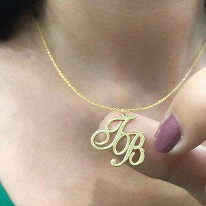 Puede incluir: Un collar de oro con un colgante de letras cursivas. El colgante es un monograma de las letras "JB".