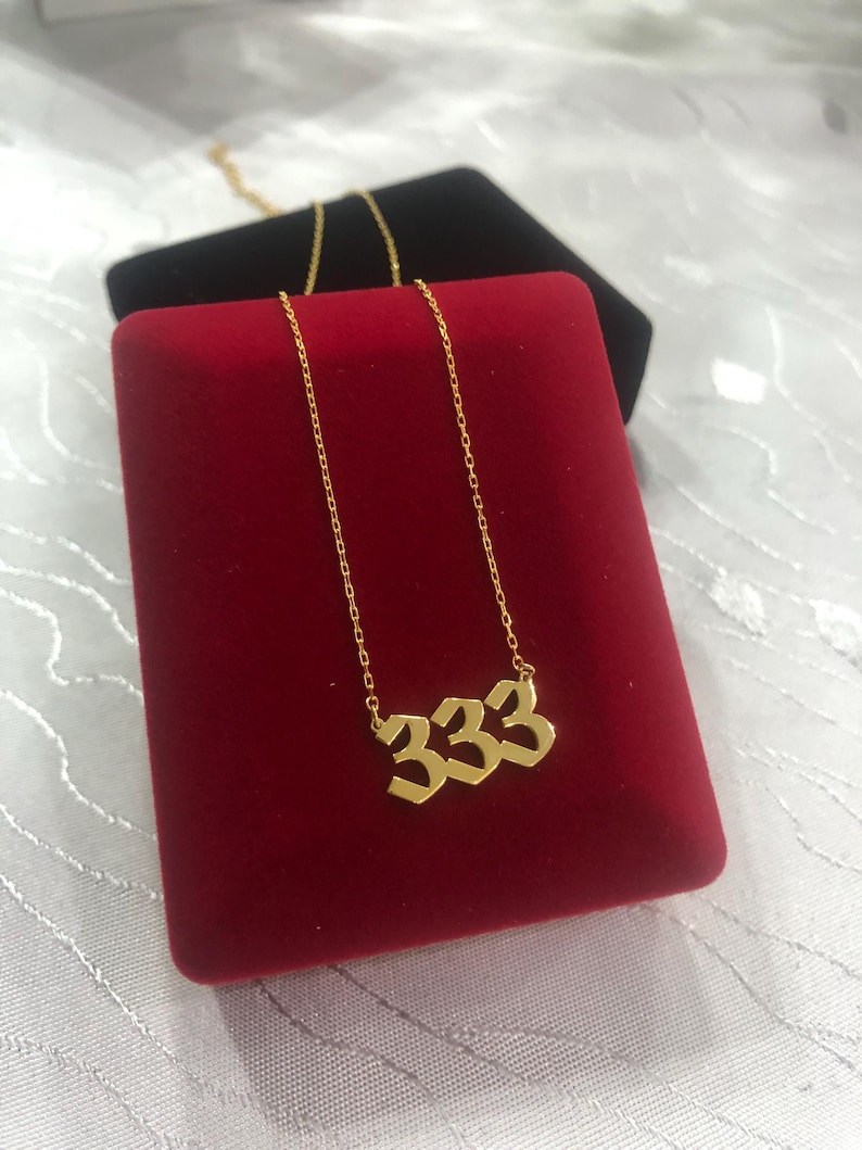 Op de afbeelding: Een gouden ketting met een hanger met het nummer 333 in een gestileerd lettertype. De ketting wordt gepresenteerd in een rode fluwelen doos.