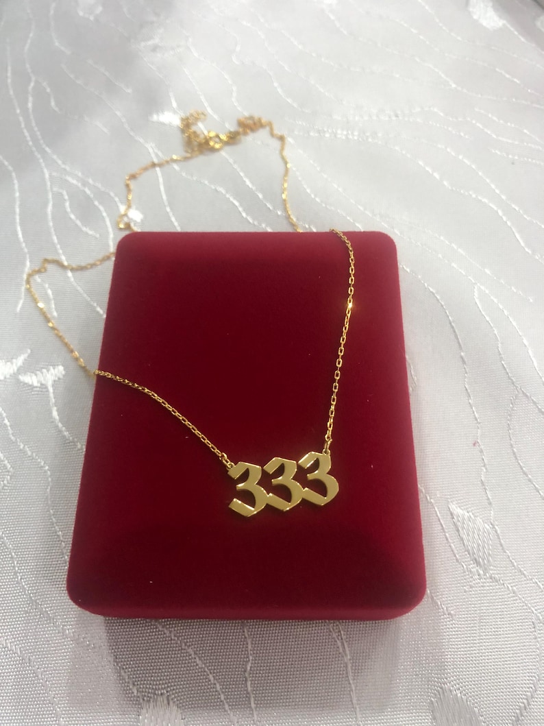 Op de afbeelding: Gouden ketting met een hanger met het nummer 333 in een gotische lettertype. De ketting wordt gepresenteerd in een rode fluwelen doos.
