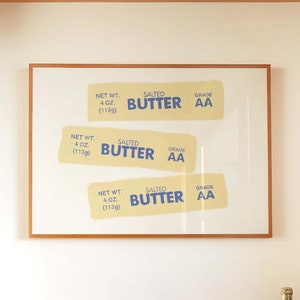May include: A framed print with three labels for salted butter, each with the net weight of 4 oz (113g) and grade AA. The print is hanging on a white wall above a marble countertop with a bowl of oranges and two bottles of wine.