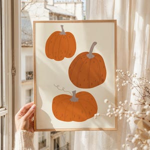 以下が含まれることがあります： クリーム色の背景に、灰色の茎が付いたオレンジ色のカボチャが3つ描かれた額入りのプリント。カボチャはさまざまなサイズと形をしています。このアートワークは、建物の見える窓の近くに展示されています。