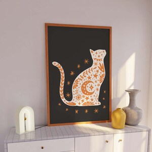 Puede incluir: Una impresión enmarcada de una silueta de gato blanco con un patrón floral naranja sobre un fondo negro. El gato está sentado con la cola enroscada alrededor de su cuerpo. El patrón floral incluye estrellas, lunas y otros elementos celestiales.