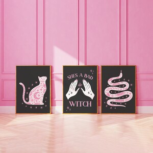May include: Three black framed prints with pink illustrations on a pink wall. The prints feature a cat, a snake, and two hands with the text "She's a bad witch".