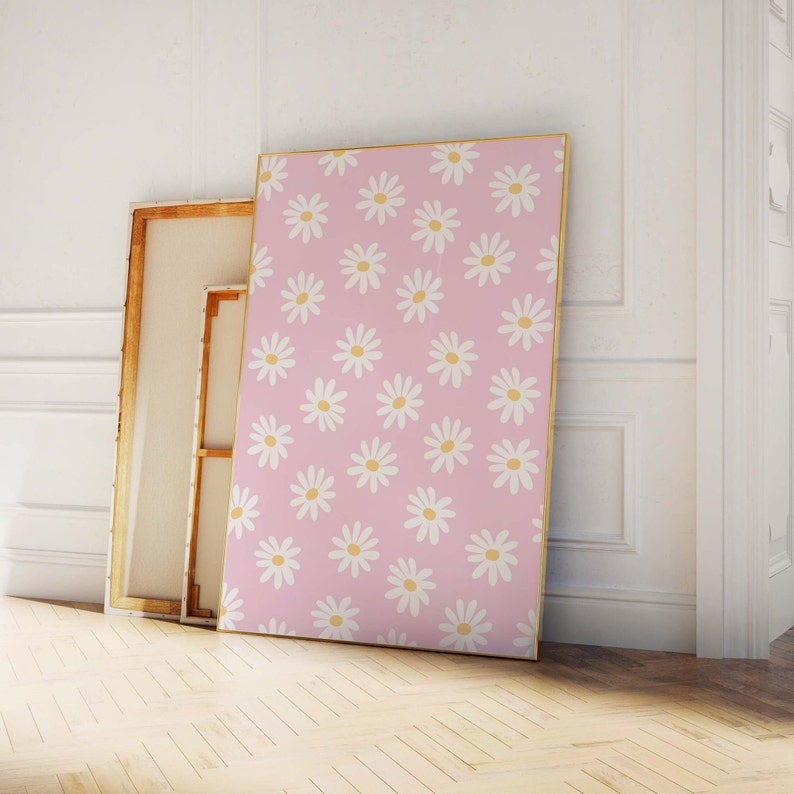 May include: A pink canvas print with a white daisy pattern. The print is framed in a gold frame and is leaning against a white wall.