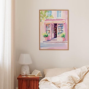 May include: A framed watercolor painting depicts a quaint bookstore with a pink facade and open doorway. The artwork features a wooden frame, potted plants, and a soft color palette of pinks, blues, and greens. The painting evokes a sense of tranquility and charm.