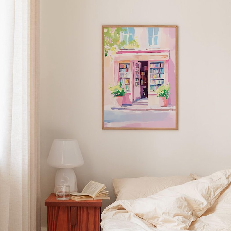 May include: A framed watercolor painting depicts a quaint bookstore with a pink facade and open doorway. The artwork features a wooden frame, potted plants, and a soft color palette of pinks, blues, and greens. The painting evokes a sense of tranquility and charm.