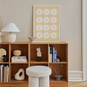 May include: A white, round, upholstered ottoman with a unique, double-leg design sits in front of a wooden bookshelf. The bookshelf is filled with books and decorative objects, including a white ceramic sculpture. A framed print with a yellow background and white daisy flowers hangs on the wall above the bookshelf.