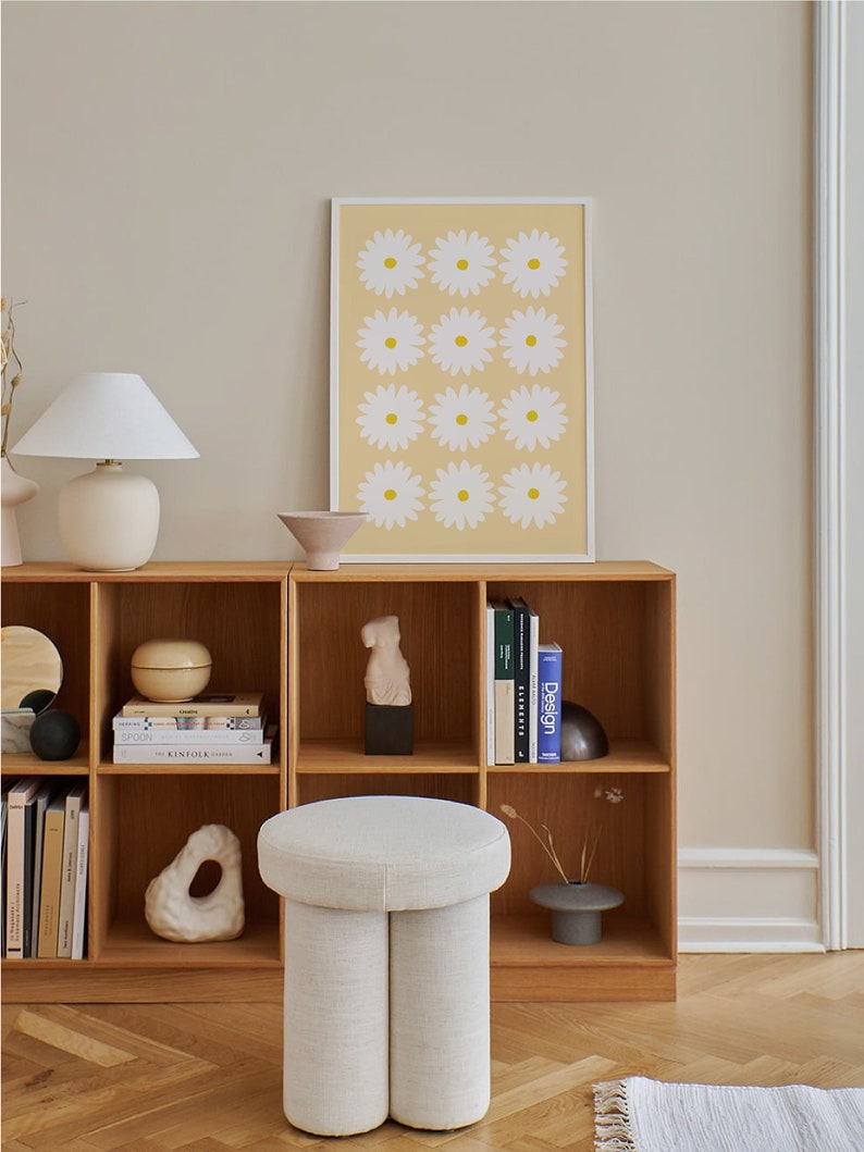 May include: A white, round, upholstered ottoman with a unique, double-leg design sits in front of a wooden bookshelf. The bookshelf is filled with books and decorative objects, including a white ceramic sculpture. A framed print with a yellow background and white daisy flowers hangs on the wall above the bookshelf.