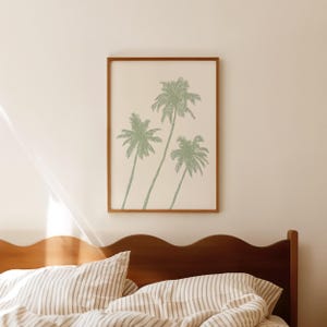 Puede incluir: Un cuadro enmarcado con tres palmeras estilizadas en un suave tono verde sobre un fondo crema. La obra de arte est&aacute; encerrada en un marco de madera marr&oacute;n c&aacute;lido. El cuadro cuelga sobre una estructura de cama de madera con ropa de cama a rayas.