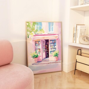 May include: A framed watercolor painting of a pink bookstore with open doors revealing bookshelves. The painting features a pink awning, green plants in pots, and blue windows. The artwork is displayed on a wooden floor next to a pink chair.