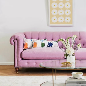 May include: A pink tufted sofa with two colourful throw pillows and a glass-top coffee table with a marble base. The coffee table is decorated with a gold leaf and a bowl of decorative items. A bouquet of white and purple flowers is in a white vase on the sofa.