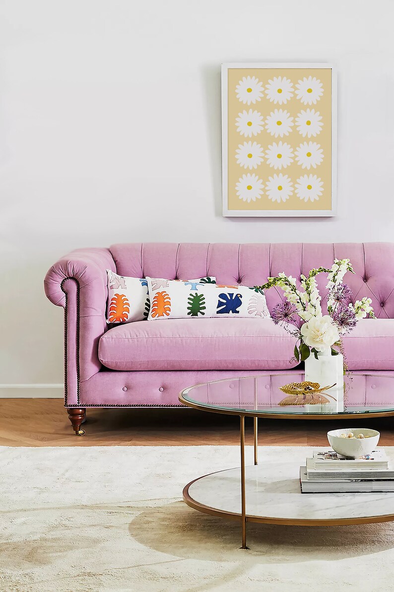May include: A pink tufted sofa with two colourful throw pillows and a glass-top coffee table with a marble base. The coffee table is decorated with a gold leaf and a bowl of decorative items. A bouquet of white and purple flowers is in a white vase on the sofa.