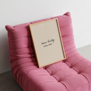 Puede incluir: Un cuadro enmarcado con el texto "how lucky are we" en escritura negra. El cuadro tiene un fondo beige y un marco de madera clara. Descansa sobre un sill&oacute;n de pana rosa.