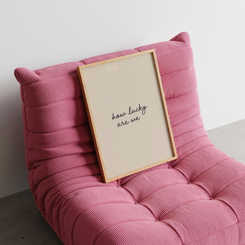 Puede incluir: Un cuadro enmarcado con el texto "how lucky are we" en escritura negra. El cuadro tiene un fondo beige y un marco de madera clara. Descansa sobre un sill&oacute;n de pana rosa.