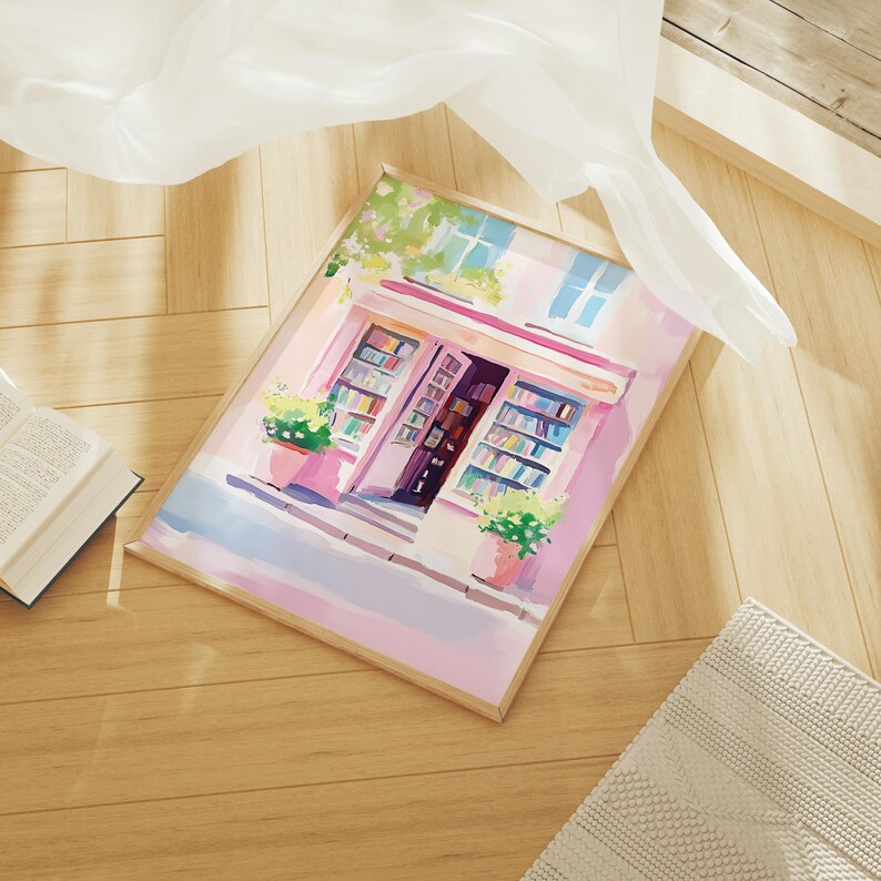 May include: Watercolor painting of a bookstore with a pink facade, open door, and colorful books. The artwork features potted plants, blue window details, and a wooden frame. The image is on a wooden surface.