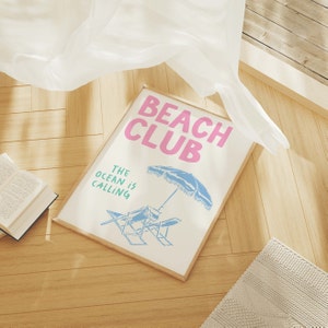 Pu&ograve; includere: Una stampa incorniciata con il testo "Beach Club" in lettere rosa e "The ocean is calling" in lettere verdi. La stampa presenta un ombrellone da spiaggia blu e bianco e due sedie a sdraio.