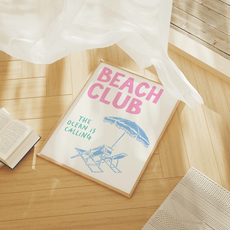 Pu&ograve; includere: Una stampa incorniciata con il testo "Beach Club" in lettere rosa e "The ocean is calling" in lettere verdi. La stampa presenta un ombrellone da spiaggia blu e bianco e due sedie a sdraio.