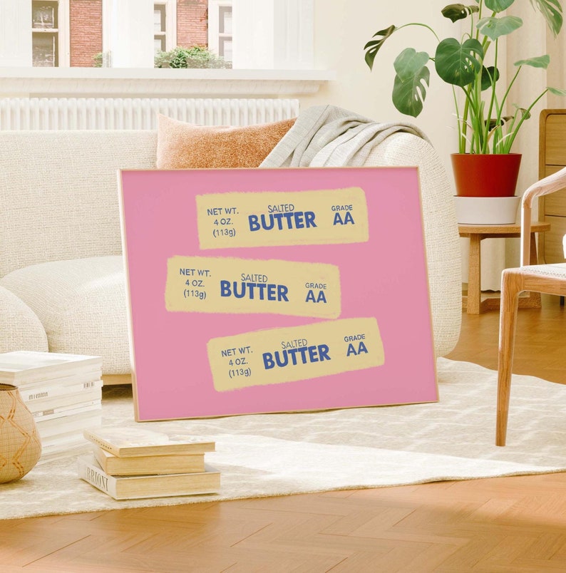 Peut inclure: Une affiche rose avec trois illustrations de plaquettes de beurre. Chaque plaquette est &eacute;tiquet&eacute;e "SALTED BUTTER" et "GRADE AA", avec un poids net de 113g. L'affiche est pr&eacute;sent&eacute;e dans un salon.