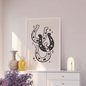 May include: A black and white print of three horseshoes with a star pattern inside each horseshoe. The print is framed in a white frame and hanging on a light pink wall.