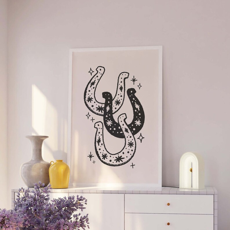 May include: A black and white print of three horseshoes with a star pattern inside each horseshoe. The print is framed in a white frame and hanging on a light pink wall.