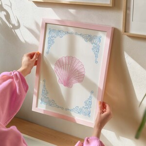 Puede incluir: Una impresi&oacute;n enmarcada en rosa con un dise&ntilde;o de concha marina azul claro y blanco. La concha marina est&aacute; en el centro de la impresi&oacute;n y est&aacute; rodeada por un borde decorativo.