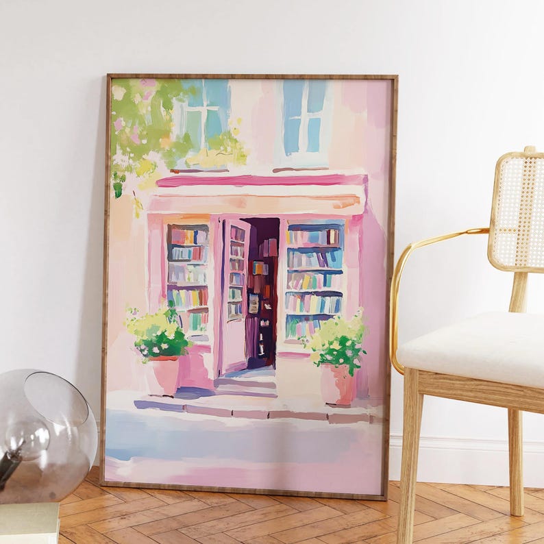 May include: A framed watercolor painting of a bookstore exterior. The building is painted in shades of pink and has open doors revealing bookshelves. Green plants in pink pots flank the entrance. The painting is in a wooden frame.