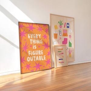 May include: Three framed art prints leaning against a white wall. The largest print is orange with pink starbursts and the text "EVERY THING is FIGURE OUTABLE." The other prints feature colorful illustrations of office supplies and objects.