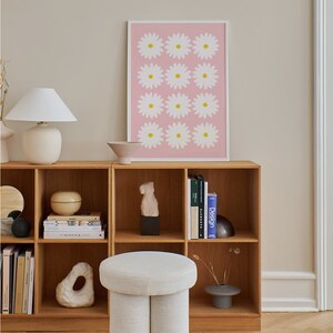 May include: A white, round, upholstered ottoman with two legs. The ottoman is in front of a wooden bookshelf with books and decorative objects. A pink framed print with white daisies is on the wall behind the bookshelf.