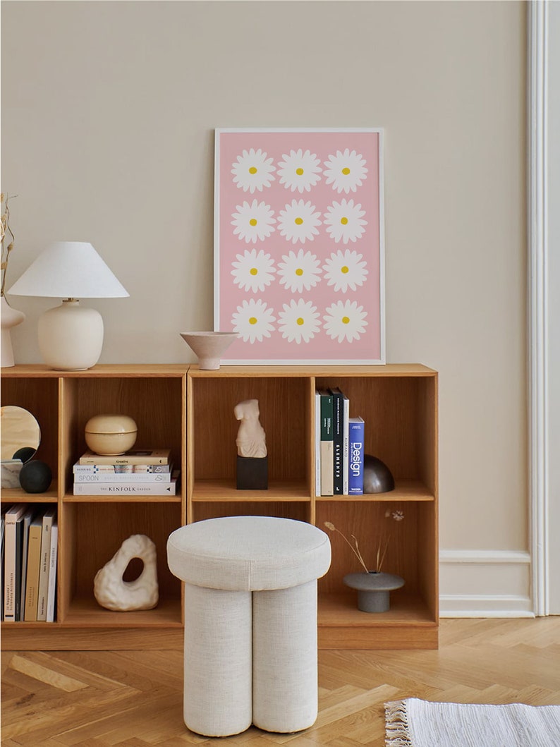 May include: A white, round, upholstered ottoman with two legs. The ottoman is in front of a wooden bookshelf with books and decorative objects. A pink framed print with white daisies is on the wall behind the bookshelf.