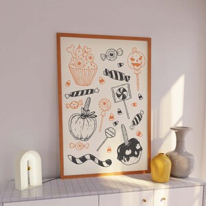 K&ouml;nnte beinhalten: Ein gerahmter Druck mit Halloween-Motiv, der verschiedene schwarze und orangefarbene Illustrationen von S&uuml;&szlig;igkeiten, Cupcakes und K&uuml;rbissen zeigt. Der Druck ist auf wei&szlig;em Hintergrund mit einem Holzrahmen.