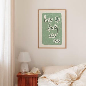 May include: A green and white print with the text "how lucky are we" and a four-leaf clover graphic. The print is framed in a light wood frame.