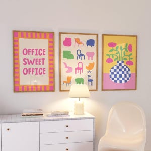May include: Three framed art prints. The left print has the text "OFFICE SWEET OFFICE" in pink on a cream background, framed with orange and pink stripes. The middle print features colorful chair illustrations. The right print shows a vase with flowers.