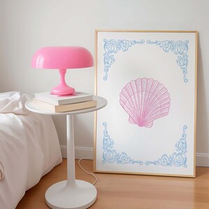 Puede incluir: Una l&aacute;mpara de mesa rosa con una pantalla blanca se encuentra sobre una mesa auxiliar blanca junto a una cama blanca. Un estampado enmarcado con una concha rosa y esquinas decorativas azules cuelga de la pared.