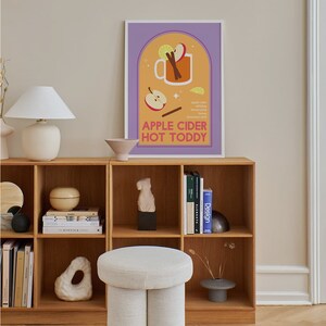May include: A colorful illustration of a mug filled with apple cider, cinnamon sticks, and a lemon wedge. The text "APPLE CIDER HOT TODDY" is below the mug. The illustration is on a poster with a purple background and a white frame. The poster is hanging on a wall above a wooden shelf with books and decorative objects.