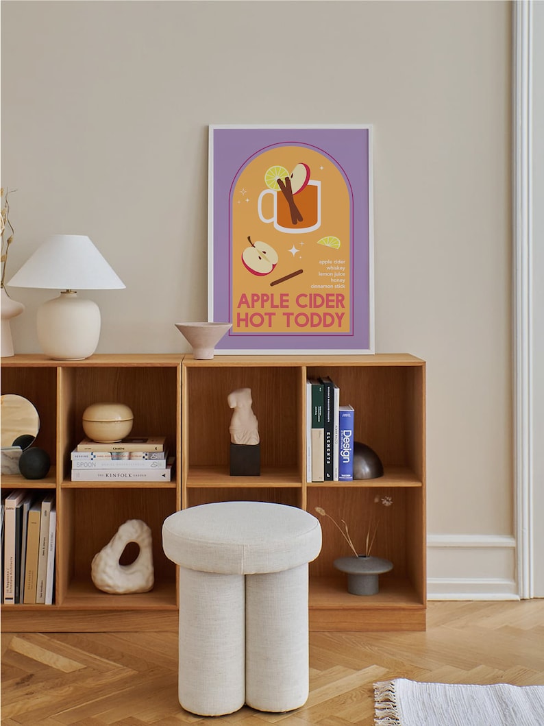 May include: A colorful illustration of a mug filled with apple cider, cinnamon sticks, and a lemon wedge. The text "APPLE CIDER HOT TODDY" is below the mug. The illustration is on a poster with a purple background and a white frame. The poster is hanging on a wall above a wooden shelf with books and decorative objects.