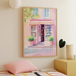 May include: A framed art print depicts a quaint bookstore facade in shades of pink and white. The open doorway reveals shelves of books, with potted plants and window details adding charm. The artwork is in a light wood frame.