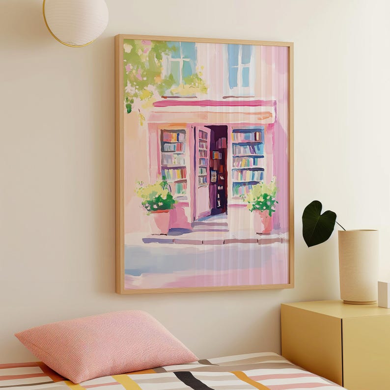 May include: A framed art print depicts a quaint bookstore facade in shades of pink and white. The open doorway reveals shelves of books, with potted plants and window details adding charm. The artwork is in a light wood frame.
