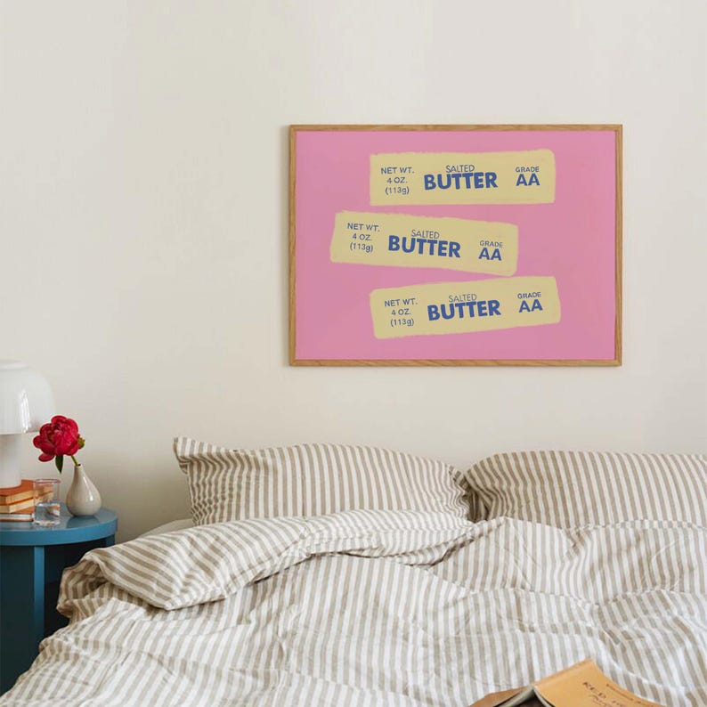 Peut inclure: Une affiche encadr&eacute;e repr&eacute;sentant trois paquets de beurre stylis&eacute;s sur fond rose. Chaque paquet porte la mention "SALTED BUTTER" et "GRADE AA", avec un poids net de 113g. L'affiche est dans un cadre en bois, au-dessus d'un lit.