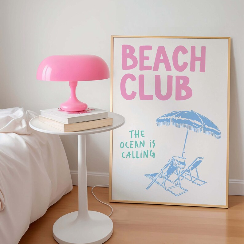 Pu&ograve; includere: Una lampada da tavolo rosa con un paralume bianco si trova su un tavolino bianco accanto a un poster incorniciato in oro con il testo "Beach Club" e "The Ocean is Calling" in lettere rosa e verdi. Il poster presenta un ombrellone da spiaggia blu e due sedie da spiaggia blu.