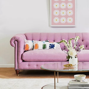 May include: A pink tufted sofa with a glass and marble coffee table. The sofa has two pillows with a colorful geometric pattern. A vase with white and purple flowers sits on the coffee table.