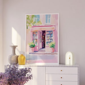 May include: A framed watercolor painting of a bookstore with an open door, filled with books. The building is painted in shades of pink and white, with green plants in pots. A vase, a yellow jug, and a lamp are on a white cabinet.