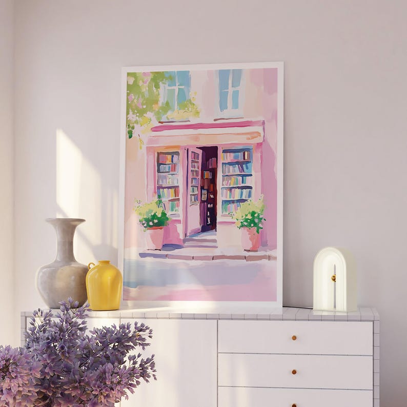 May include: A framed watercolor painting of a bookstore with an open door, filled with books. The building is painted in shades of pink and white, with green plants in pots. A vase, a yellow jug, and a lamp are on a white cabinet.