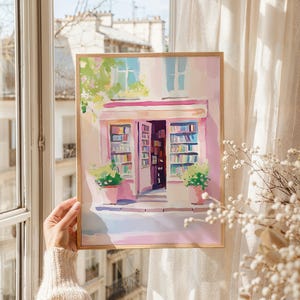 May include: Watercolor painting of a Parisian bookstore with a pink facade, open doors, and shelves filled with books. The artwork features a wooden frame and potted plants, evoking a charming, vintage aesthetic. The scene is bathed in soft, natural light.