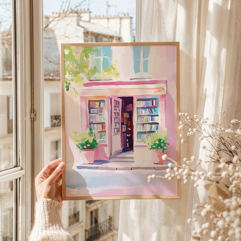 May include: Watercolor painting of a Parisian bookstore with a pink facade, open doors, and shelves filled with books. The artwork features a wooden frame and potted plants, evoking a charming, vintage aesthetic. The scene is bathed in soft, natural light.