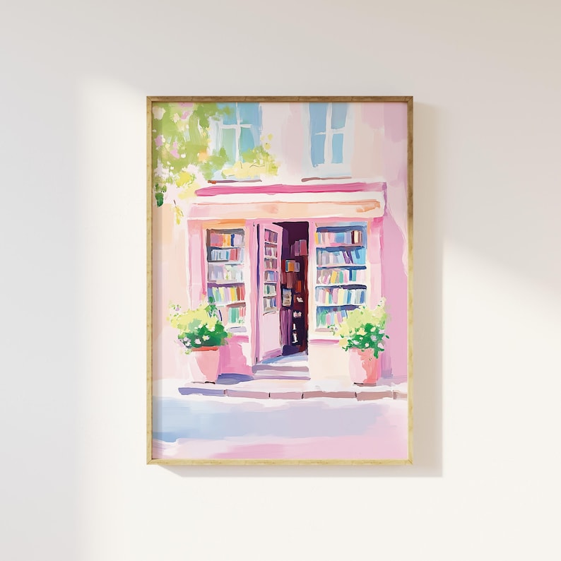 May include: A framed watercolor painting of a pink bookstore with open doors, displaying shelves of books. The storefront has a striped awning and potted plants. The artwork is in a gold frame and set against a white wall.
