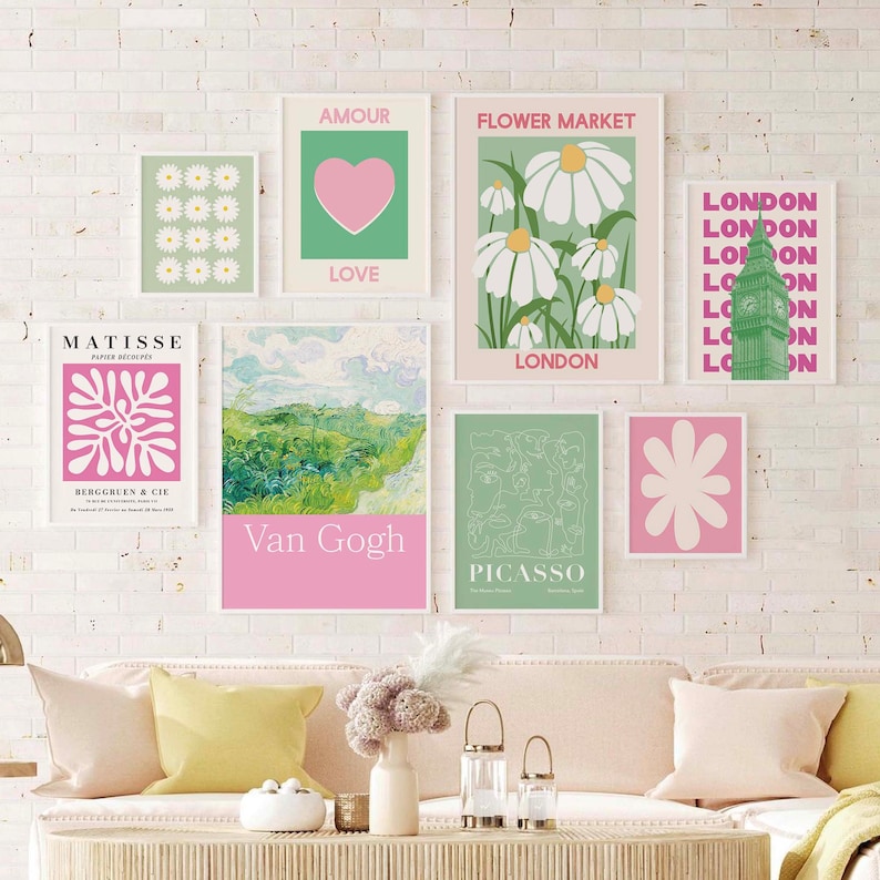 May include: A collection of eight framed art prints in a pink and green colour scheme. The prints feature various designs, including a heart, flowers, a clock tower, and abstract shapes. The prints are arranged on a white brick wall above a light pink sofa.