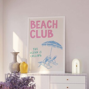 Pu&ograve; includere: Un poster beige con il testo "Beach Club" in rosa e "The Ocean is Calling" in verde. Il poster presenta un ombrellone da spiaggia blu e due sdraio blu su una spiaggia bianca.
