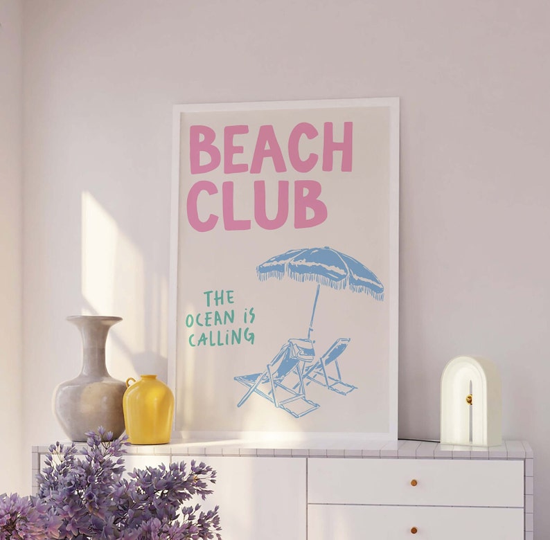 Pu&ograve; includere: Un poster beige con il testo "Beach Club" in rosa e "The Ocean is Calling" in verde. Il poster presenta un ombrellone da spiaggia blu e due sdraio blu su una spiaggia bianca.