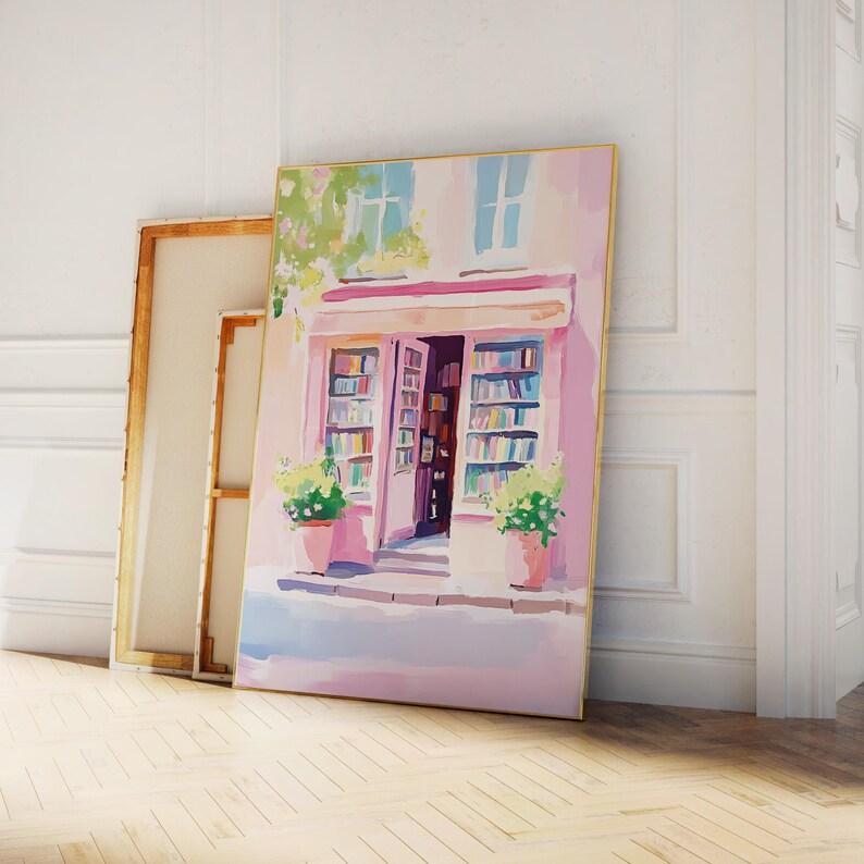 May include: A framed painting depicts a quaint bookstore facade in soft pinks and blues. The open doorway reveals shelves of books. Two potted plants flank the entrance. A second, unframed canvas leans against the wall, suggesting an art studio or gallery setting.