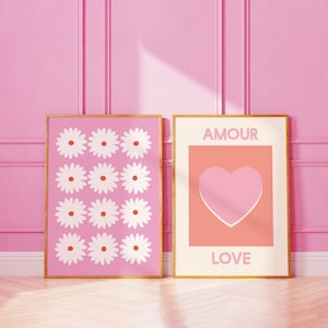 May include: Two framed prints with a pink background. The print on the left features a pattern of white flowers with red centers. The print on the right features a pink heart on a peach background with the words "AMOUR" and "LOVE" in white.